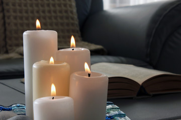 Burning candles, pebbles, glass tray, open book in gray sofa with blanket background. Interior decoration, living room.