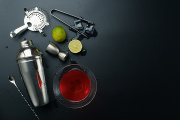 Cosmopolitan cocktail and bar equipments, stainless steel cocktail shaker and jigger, bar spoon with strainer, the lemons and ice tongs with ice cubes on the table