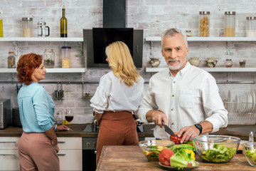 handsome man preparing salad for dinner at home, women talking with wine