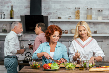 smiling attractive women preparing salad for dinner, men talking with wine at home