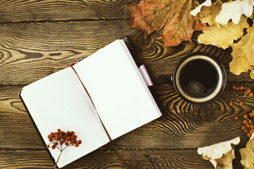Top view of the diary, a cup of hot coffee, autumn oak leaves and rowan berries on a wooden background. Copy space. Cozy. Autumn concept.