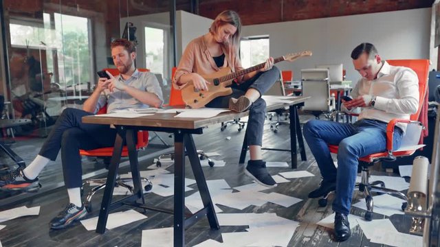 Business group of young people are bored in the office without work after a failure of business meeting. Woman with guitar setting on table, two mens playing with a phones, paper is scattered in floor