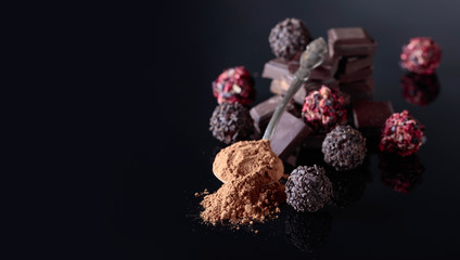 Chocolate candies, broken chocolate pieces and spoon with cocoa powder on a black reflective background.