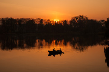Young couple in a boat sailing on a lake at sunset. Silhouettes of romantic couple sailing in a boat against the backdrop of the orange sunset.