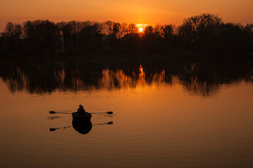 Naklejka premium Young couple in a boat sailing on a lake at sunset. Silhouettes of romantic couple sailing in a boat against the backdrop of the orange sunset.