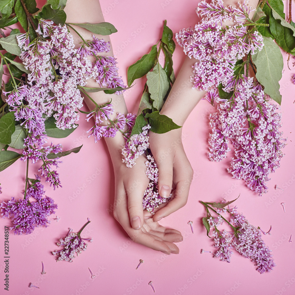 Wall mural Fashion art hands natural cosmetics women, bright purple lilac flowers in hand with bright contrast makeup, hand care. Creative beauty photo of a girl sitting at table on contrasting pink background