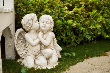 2 Cupids are embraced in a garden near a bench.