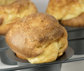 Homemade popover, which is a puffed, airy, and eggy hollow roll, is fresh from the oven in a dedicated metal pan with two more blurred popovers in the gray background.