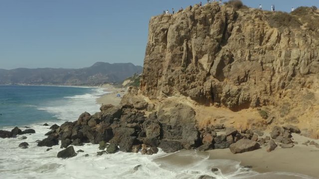 Aerial pullback over the waves and rocks of a cliff on the beach.