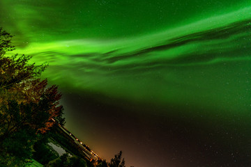 inclined earth and bright green aurora lights almost on the whole sky over tree tops in Sweden, river, city lights and lake, clear skies with a lot of stars, pine trees, autumn colored leaves on trees