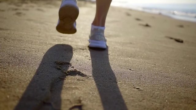 View from behind of a woman walking on wtd sand on a beach in white sneakers, warm summer day.