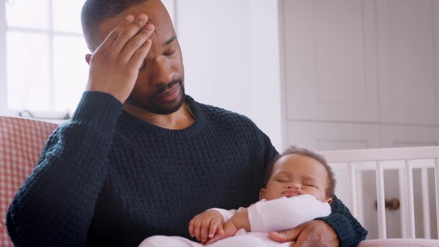 Stressed New Father Sitting In Chair Holding Sleeping Baby Girl In Nursery At Home