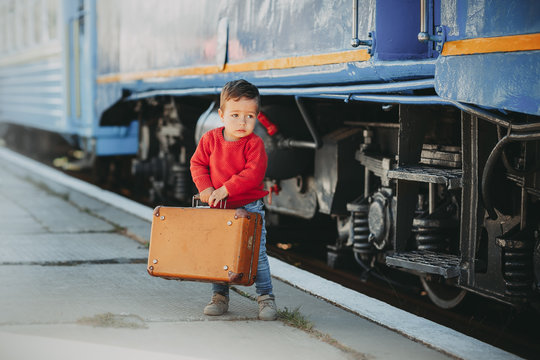 Adorable little kid boy dressed in red sweater on a railway station near train with retro old  brown suitcase. Ready for vacation. Young traveler on the platform.