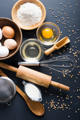 Baking ingredients. Bowl, eggs, flour, eggbeater, rolling pin and eggshells on black chalkboard from above.