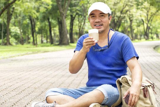 People man holding coffee cup or disposable cup with a black plastic cap and backpack sitting in the garden green nature background. Concept happy man with leisure and drink coffee.