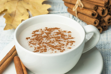 white cup of salep milky hot drink of Turkey with cinnamon powder and sticks healthy spice and autumn winter leaf on white rustic vintage wooden table. Sahlep background
