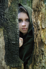 young beautiful elf in a cloak hides behind a trunk of a burnt tree and looks out into the gap
