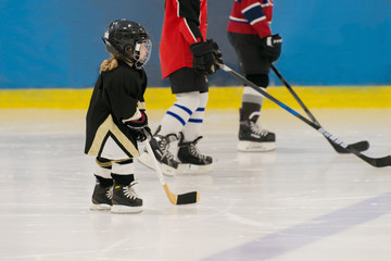 Fototapeta premium The little cute hockey girl is on the ice wearing in full equipment: hockey helmet, gloves, stick, skates. Figures of two teenagers hockey players are on the background.