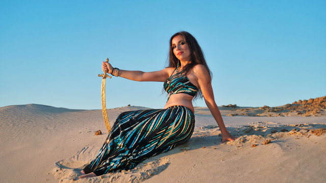 Oriental Beauty dancing belly dance with scimitar in desert. Girl is sitting on sand. Dangerous houri and heavenly pleasures. Portrait on background of golden sand dunes at sunset