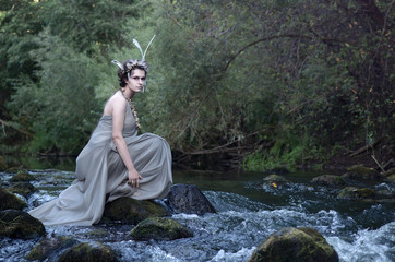beautiful young woman in gray silk dress, headdress of feathers and necklace of sea snails, forest nymph, sitting on a river stone and resting