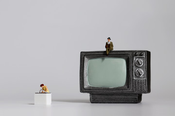 Miniature man sitting on miniature television and a miniature woman doing household chores.