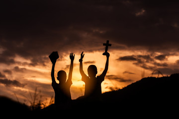 Two children worship God by holding christian Cross and Bible with light sunset background,christian concept.