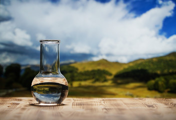 Clean water in a glass laboratory flask on wooden table on mountain background. Ecological concept,...