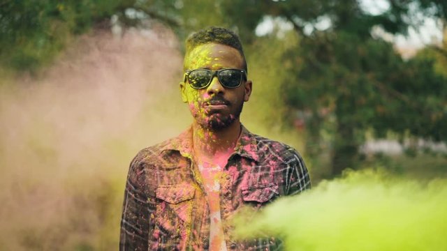 Portrait of excited African American man celebrating Holi festival standing outdoors in park and laughing while people are throwing powder paint at him.