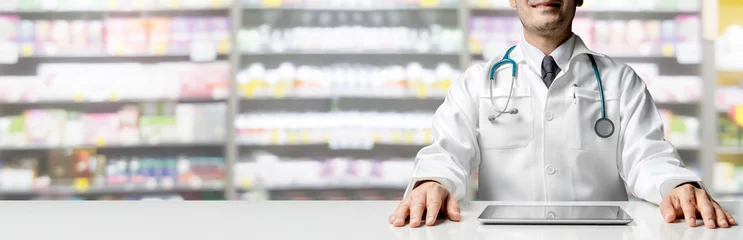 Wall murals Pharmacy Male pharmacist sitting at table with tablet computer in pharmacy office. Medical healthcare staff and drugstore business.