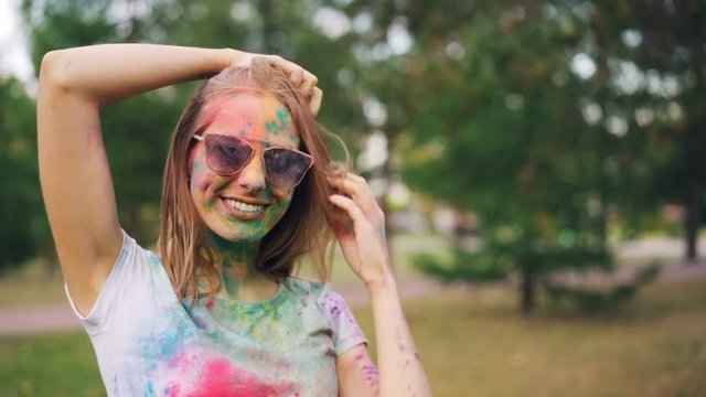 Portrait of attractive young woman covered with colorful paint at Holi festival smiling, looking at camera and touching her hair standing in park outdoors.