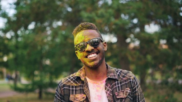Slow motion portrait of bearded African American guy in sunglasses, face and hair covered with powder paint at Holi festival. Man is looking at camera and smiling.