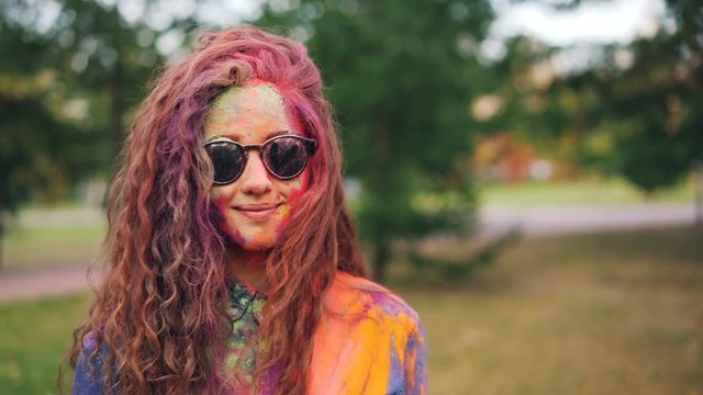 Slow motion portrait of happy girl with long curly hair covered with multicolor paint powder at Holi festival standing outdoors and smiling wearing sunglasses.