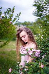 a young teenager girl with long curled hair dressed in pink standing at a blooming dog-rose bush