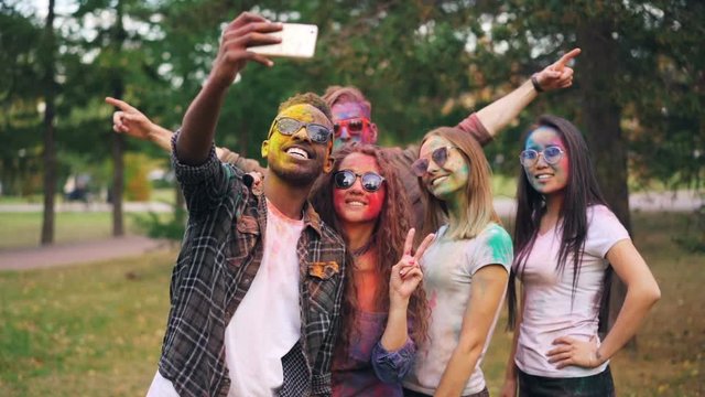 Slow motion of happy students multiethnic group with coloured faces and hair taking selfie in park using smartphone camera and having fun at Holi festival.