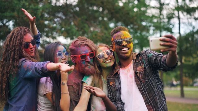 Slow motion of carefree students with coloured faces taking selfie with smartphone standing outdoors in park and posing for camera. Holi festival and photo concept.