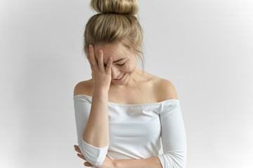 Gorgeous young female wearing off shoulder white top keeping hand on her face, covering one half, feeling shy. Charming cute girl with hair knot looking down and laughing at awkward moment