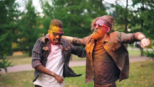 Two bearded guys with colorful faces covered with paint are dancing on lawn in park jumping and smiling enjoying party. Men with cool hairstyle are wearing sunglasses.