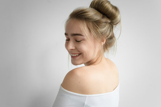 Charming girl with hair bun and nude back posing isolated at white studio wall, closing eyes and smiling broadly. Picture of attractive young Caucasian woman with messy hairstyle enjoying good day