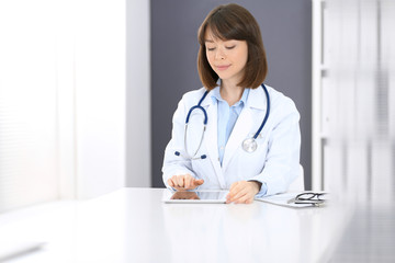 Female doctor typing on laptop computer while sitting at the white table in hospital office. Physician at work. Medicine and healthcare concept