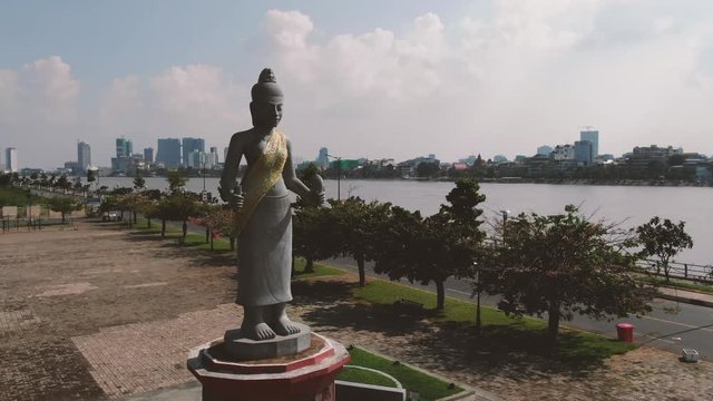 Aerial circle round a large Angkorian style statue standing on a pedestal in Cambodia, Southeast Asia. Modern city and river view can be seen in backrgound.