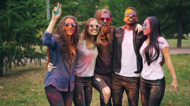 Slow motion portrait of excited young women and men in dirty clothing jumping together on lawn in park with colorful faces and hands having fun at party.