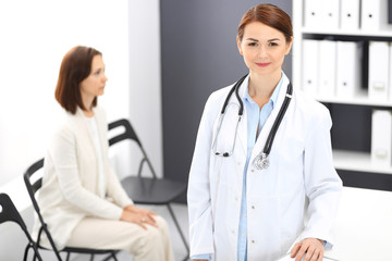 Doctor woman at work. Portrait of female physician cheerful smiling while standing near reception desk at clinic or emergency hospital. Patient woman sitting at the background. Medicine concept