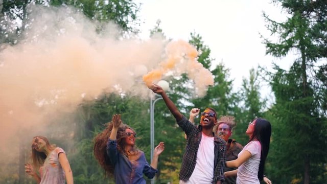 Slow motion of girls and guys dancing and jumping with smoke flares at outdoor party, skin and clothes are covered with bright paint. Emotions and holidays concept.