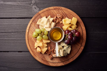 Cheese platter: Parmesan, cheddar, gouda, gorgonzola, brie and other with walnuts, olives and honey on wooden board on black background