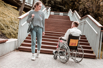 Unusual problem. Serious young woman passing the invalid man sitting in the wheelchair in front of the stairs - obrazy, fototapety, plakaty