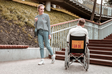 serious glance. Serious young woman walking and looking at the disabled man sitting in the wheelchair in front of high stairs