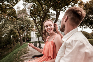 Fototapeta premium Touching hair. Disabled girl sitting in the wheelchair and holding silver balloon while caring boyfriend gently touching her hair