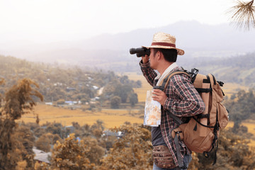 Young man with backpack and holding a binoculars and map paper is standing top of a mountain.