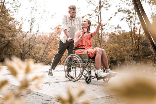 Looking excited. Cheerful emotional disabled girl sitting in the wheelchair and pointing to the distance while showing beautiful view to her smiling boyfriend