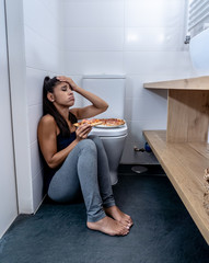 Young beautiful bulimic woman sitting on the bathroom floor eating pizza looking guilty